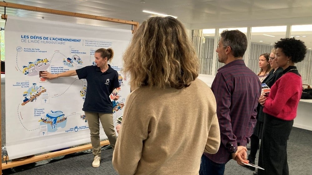Cinq personnes debout dans une pièce assistent à une présentation par une sixième qui leur montre des dessins sur un panneau.