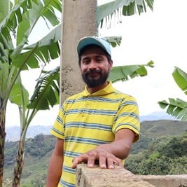 Un homme barbu coiffé d'une casquette pose en extérieur et regarde l'objectif en souriant.